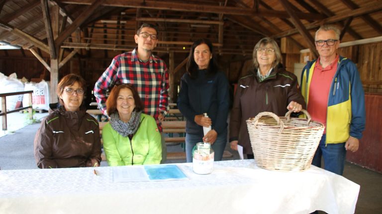 Five adults are smiling for a photo in a rustic setting. A man stands behind a table with a jar and a basket. Two women are seated at the table with books and a pen.