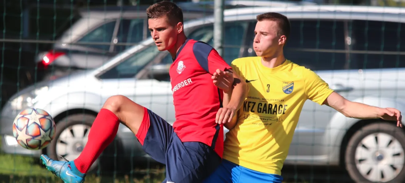 Zwei Fußballspieler in roten und gelben Trikots, einer mit erhobenem Fuß und der andere mit erhobenem Arm. Dahinter ist ein Zaun und ein verschwommener Hintergrund eines geparkten Autos.