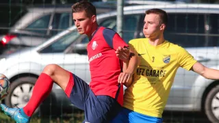 Zwei Fußballspieler in roten und gelben Trikots, einer mit erhobenem Fuß und der andere mit erhobenem Arm. Dahinter ist ein Zaun und ein verschwommener Hintergrund eines geparkten Autos.