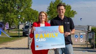 Zwei Menschen stehen lächelnd draußen und halten ein blaues Schild mit der Aufschrift Burgenland Family Card. Die Frau trägt ein rotes Outfit, der Mann ein schwarzes Hemd. Hinter ihnen steht ein Metalltor mit mehreren Schildern.