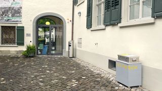 Ein Eingang mit Glastüren und grünen Fensterläden an einem weißen Gebäude. Ein Mülleimer und ein Müllkasten befinden sich an der Seite. Eine Topfpflanze steht links.