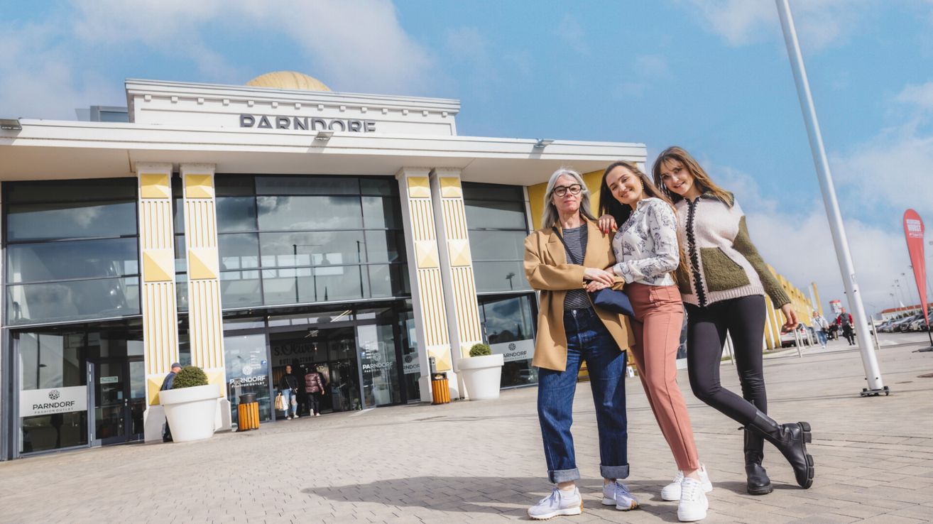 Drei Frauen posieren vor einem modernen Gebäude mit einem Schild, das Parndorf sagt. Sie stehen vor großen Glasfenstern, wobei zwei Frauen Brillen und Turnschuhe tragen.