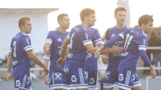 Fünf männliche Fußballspieler in blauen Trikots stehen eng beieinander und feiern, wahrscheinlich nach einem Spiel. Ein Spieler trägt ein Trikot mit der Nummer 6, und alle haben Recycling-Logos auf ihren Shorts.