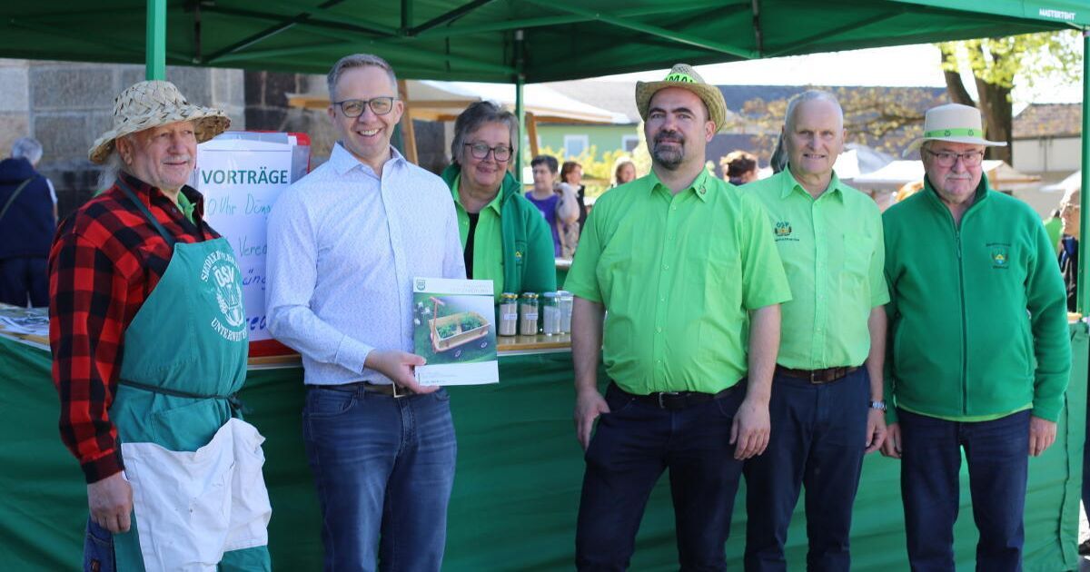 Vier Menschen stehen unter einem Zelt auf einem Markt. Ein Mann hält ein Buch und lächelt. Die anderen tragen grüne Hemden und einer trägt einen Strohhut. Dahinter steht ein Schild mit der Aufschrift Vorträge.