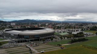 Luftaufnahme eines großen Stadions mit Feld, geparkten Autos, Gebäuden und Bäumen. In der Ferne sind Berge unter einem bewölkten Himmel zu sehen.