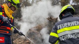 Zwei Feuerwehrleute sprühen Wasser auf ein rauchiges Feuer. Einer hält einen Schlauch und der andere trägt einen gelben Helm.