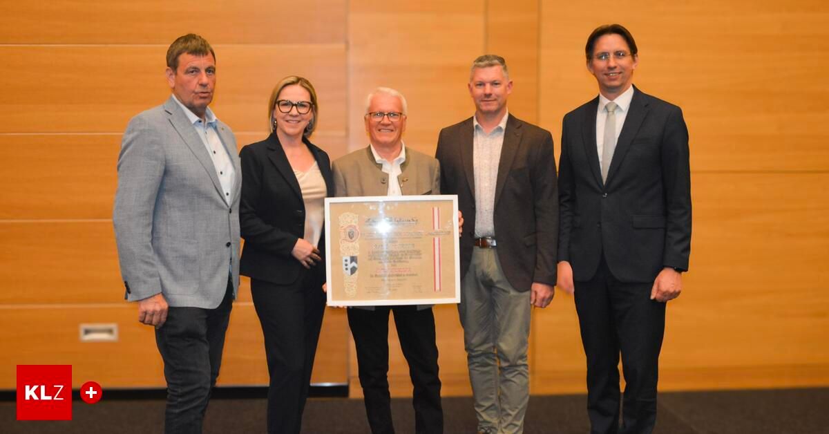 Fünf Personen stehen eng beieinander und halten eine Plakette mit Text und Logos. Sie sind in formeller Kleidung gekleidet, möglicherweise bei einer Preisverleihung.