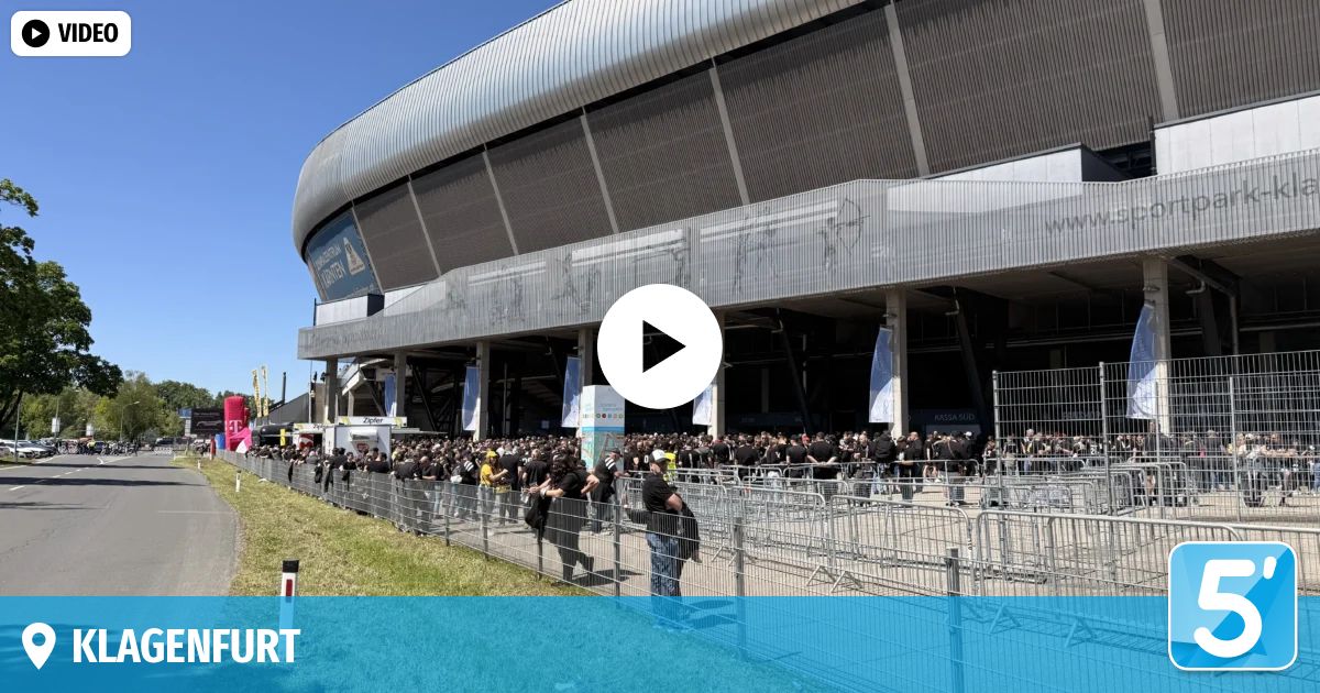 Eine große Menschenmenge in schwarzen T-Shirts wartet vor einem modernen Stadion. Der Eingang ist durch Metallzäune blockiert. Ein Video-Play-Button ist überlagert.
