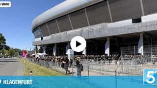 Eine große Menschenmenge in schwarzen T-Shirts wartet vor einem modernen Stadion. Der Eingang ist durch Metallzäune blockiert. Ein Video-Play-Button ist überlagert.