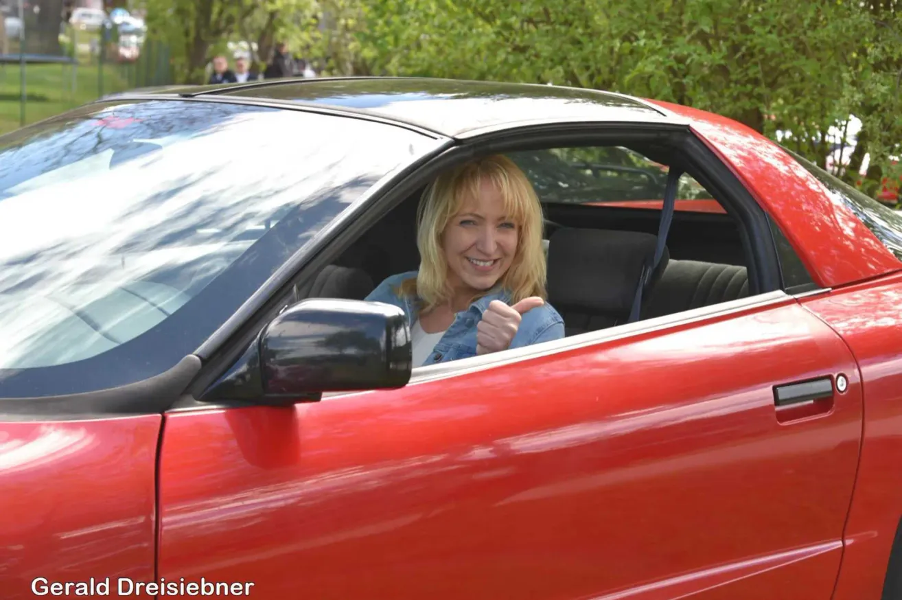 Eine Frau in einem roten Auto gibt ein Daumen hoch. Sie sitzt im Fahrersitz und lächelt. Bäume sind im Hintergrund sichtbar.