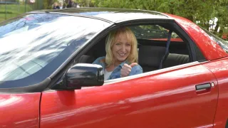 Eine Frau in einem roten Auto gibt ein Daumen hoch. Sie sitzt im Fahrersitz und lächelt. Bäume sind im Hintergrund sichtbar.