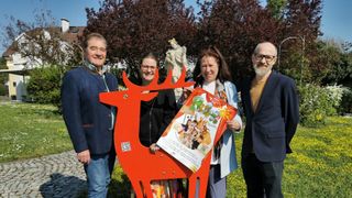Vier Personen stehen hinter einer großen roten Hirschskulptur in einem Park und halten ein Plakat für eine Veranstaltung. Der Mann links lächelt, während die Frau in der Mitte in die Kamera schaut. Die Frau rechts hält das Plakat und der Mann rechts trägt eine Brille. Dahinter sind eine Statue und ein Baum zu sehen.