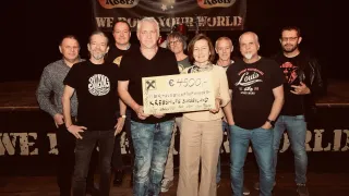 Gruppe von Menschen in schwarzen T-Shirts posiert mit einer Frau, die ein Schild mit der Aufschrift '4500,-' hält. Ein Banner mit den Worten 'WE ROCK YOUR WORLD' ist im Hintergrund.