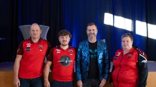 Vier Männer stehen zusammen. Der Mann auf der linken Seite trägt ein rotes Poloshirt mit Logo. Der Mann neben ihm trägt einen blauen Blazer. Der Mann auf der rechten Seite trägt ein rotes Shirt. Sie stehen vor einem blauen Vorhang.