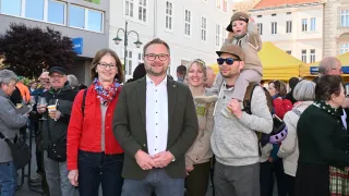 Eine Gruppe von Erwachsenen und Kindern steht vor einem Gebäude, ein Mann trägt ein Kind auf seinen Schultern. Die Frau links trägt eine rote Jacke. Der Mann in der Mitte hat ein Lächeln im Gesicht.