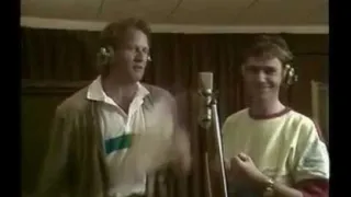 Zwei Männer stehen nebeneinander in einem Aufnahmestudio, einer trägt Kopfhörer und der andere eine Uhr. Sie haben beide Mikrofone vor sich und scheinen zu aufnehmen.