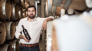 Ein Mann in einem weißen Hemd und einem braunen Gürtel steht in einem Weinkeller und hält eine Flasche Argo-Wein. Hinter ihm befinden sich große Holzfässer.