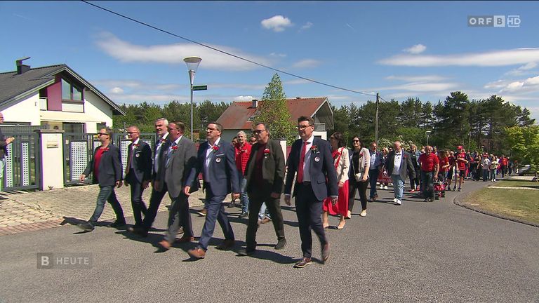 Eine Gruppe formell gekleideter Männer und Frauen geht unter einem klaren blauen Himmel eine Straße entlang, mit ein paar Häusern im Hintergrund.