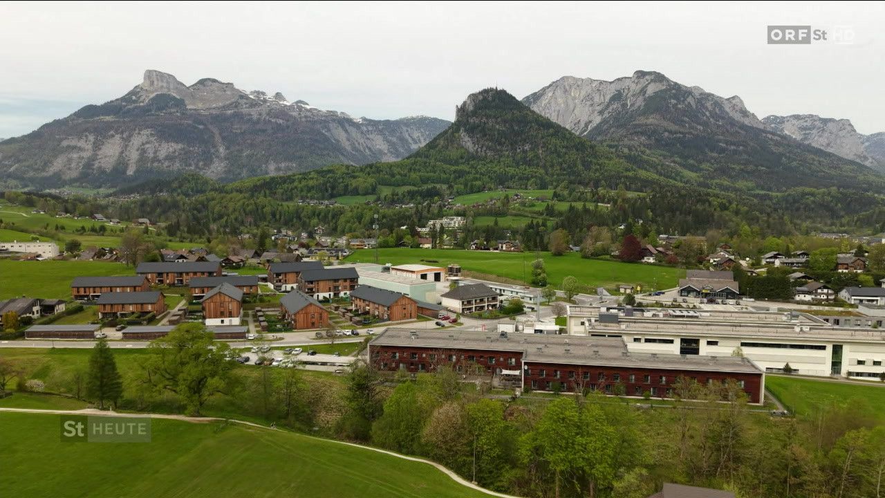 Luftaufnahme eines Dorfes in einem Gebirgstal mit mehreren modernen Gebäuden, üppigen grünen Feldern und einem klaren blauen Himmel.