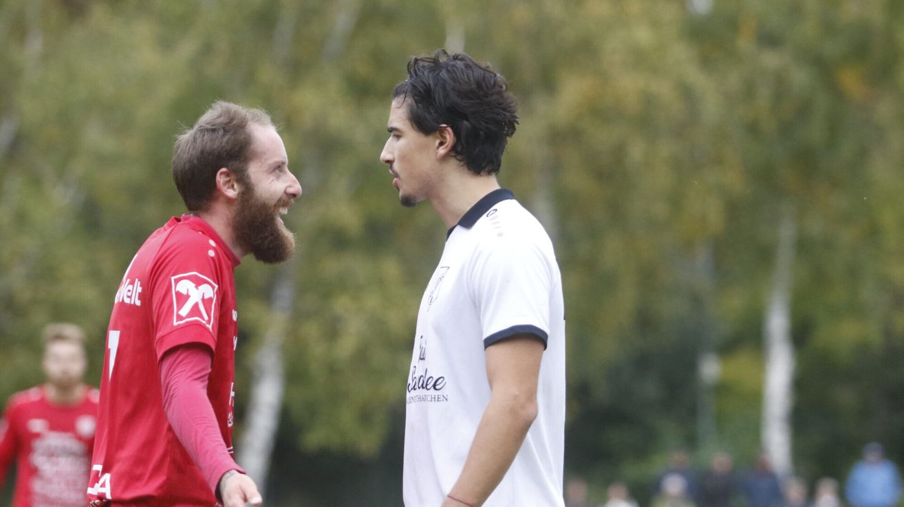 Zwei Männer in Sporttrikots stehen sich im Freien gegenüber. Einer hat einen Bart, der andere trägt ein weißes Shirt mit blauem Saum. Dahinter vermuten verschwommene Bäume und Menschen ein Sportereignis.