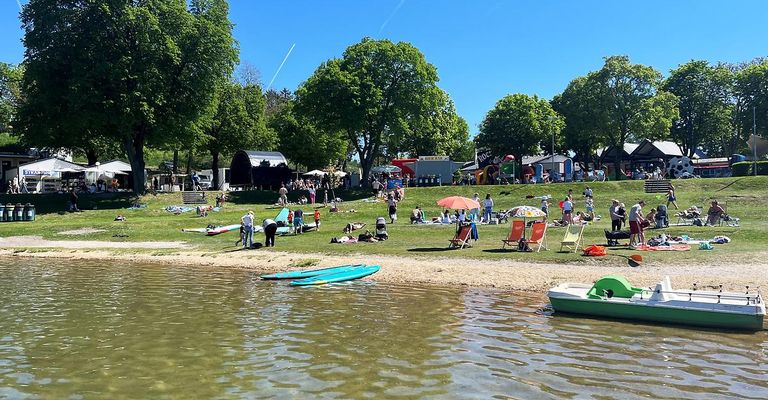 Ein sonniger Tag am See mit vielen Menschen, die sich entspannen und die Aussicht genießen, einige sonnenbaden, während andere auf Stühlen mit Sonnenschirmen sitzen. Bäume und eine Bühne sind ebenfalls in der Szene zu sehen.
