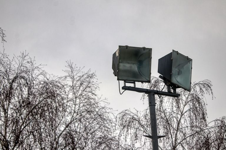 Two spotlights are mounted on a pole against a backdrop of bare trees and a cloudy sky.