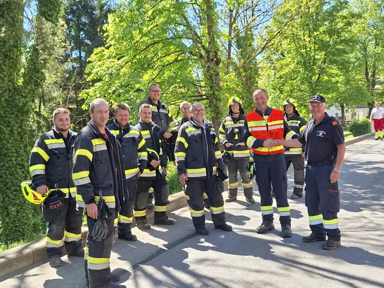 A group of firefighters in uniform stands in a line, smiling and shaking hands. The setting is outdoors with trees in the background.