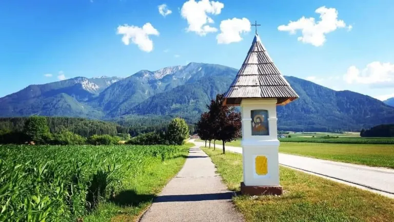 Eine kleine Kapelle mit einem Kreuz steht neben einer Straße mit Bergen im Hintergrund.