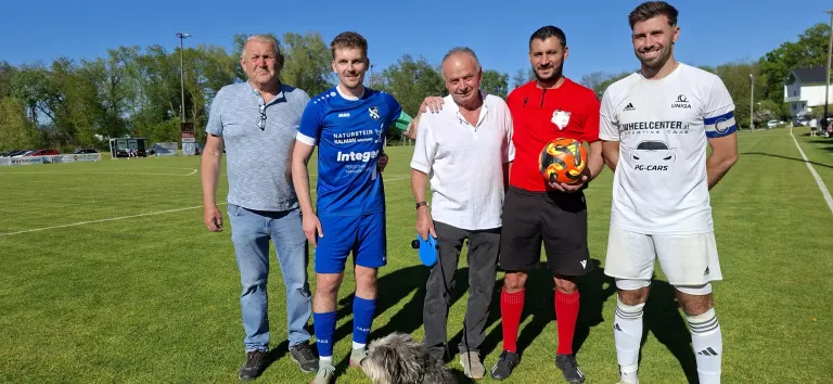 Four men in different colored shirts stand on a soccer field with a dog, one man holds a soccer ball.