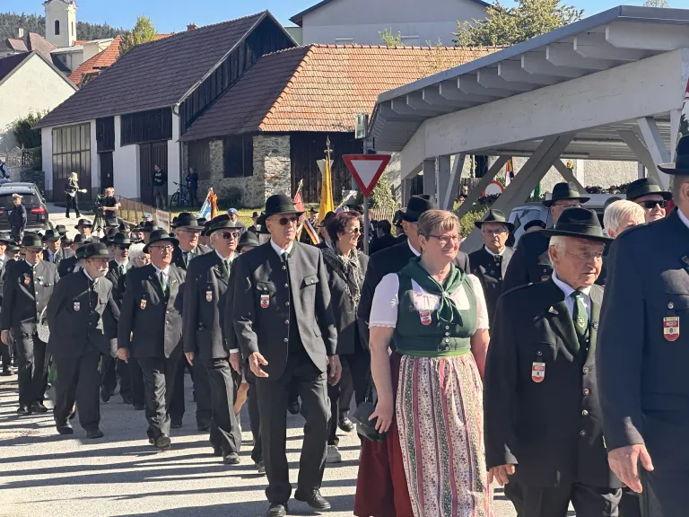 Eine Gruppe von Menschen in formeller Kleidung, einschließlich Hüten und grünen Krawatten, marschiert bei einem Umzug. Eine Frau in einer grünen Weste und einem bunten Rock sticht hervor. Hinter ihnen sind Gebäude mit gefliesten Dächern und Flaggen zu sehen.