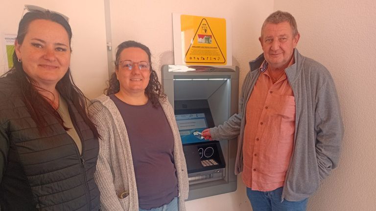 A woman and a man stand beside an ATM. The woman holds a card, and the man seems to be giving her instructions. Behind them is a yellow sign with symbols.