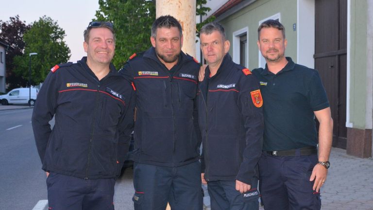 Vier Männer in Uniformen posieren für ein Foto vor einem Gebäude. Sie tragen dunkle Jacken mit verschiedenen Aufnähern und roten Streifen. Bäume und eine Straßenlaterne sind im Hintergrund zu sehen.
