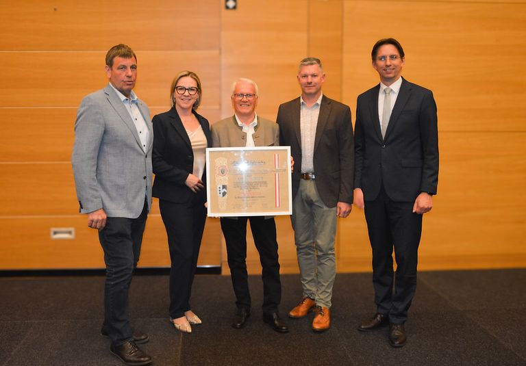 Five people stand in a room, posing for a photo. A man in the middle holds a certificate. The person on the left wears a gray blazer. The person in the middle wears glasses.
