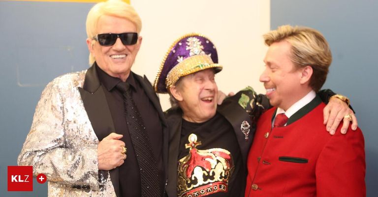 Three men in formal attire pose together. The man on the left wears a sequined jacket. The middle man wears a hat with a crown and gold embroidery. The man on the right wears a red jacket with a green collar.