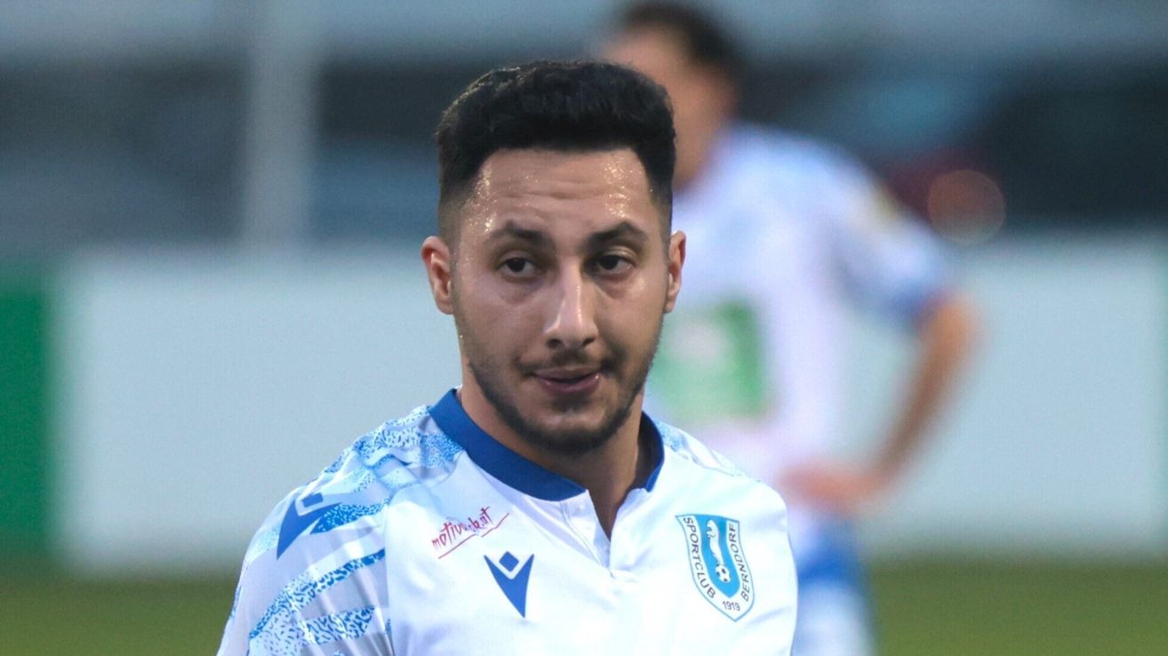 Ein Fußballer in einem weißen und blauen Trikot mit Logo auf der Brust schaut nach vorne. Dahinter ist ein weiterer Spieler in Weiß verschwommen.