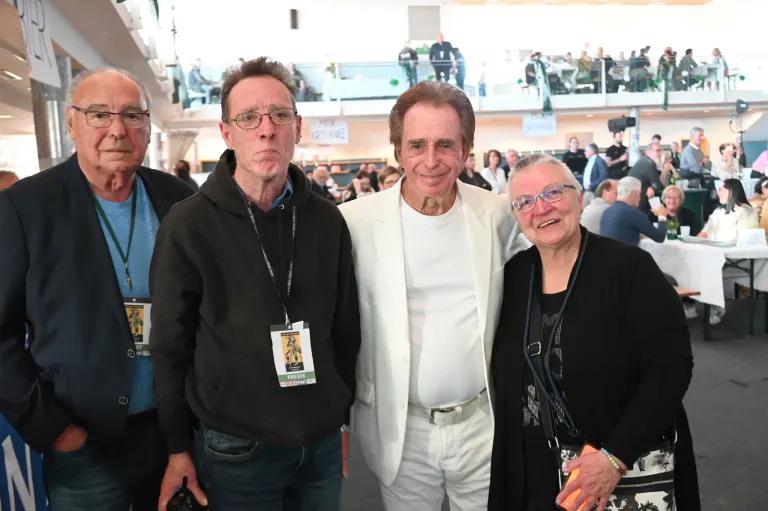 Three people stand together for a photo, with one wearing a black hoodie and another in a white suit. A woman in glasses stands beside them. Behind them, there are people sitting on chairs.