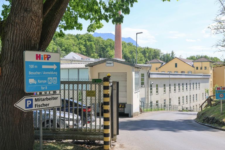 Industriegebiet mit Gebäuden und einer Fabrik im Hintergrund. Ein Schild mit 'HiPP' und einem Pfeil nach links. Ein weißer Wagen ist geparkt und ein Gebäude mit einem Rampenschild.