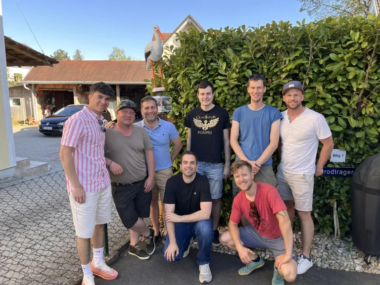 Seven men pose for a photo in front of a hedge, with a stork statue and a house in the background. They are dressed casually, smiling, and appear to be friends.
