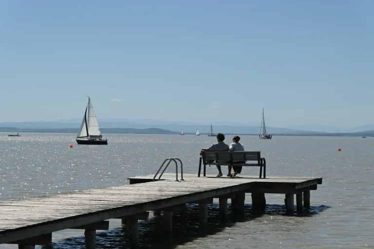 Two people sit on a bench on a wooden pier, overlooking a body of water with sailboats in the distance. The sky is clear with a few clouds.