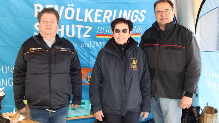 Drei Personen stehen vor einem blauen Banner mit weißen Texten. Die Frau in der Mitte trägt eine Sonnenbrille und eine Jacke mit gelbem Logo. Die Männer tragen schwarze Jacken.