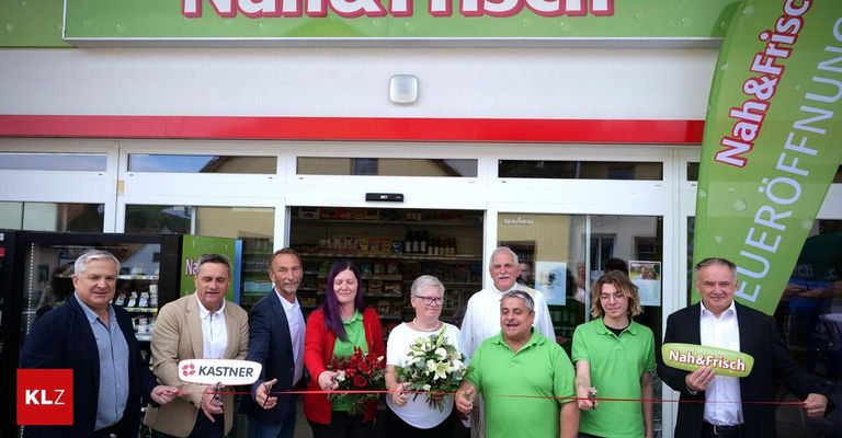 Several people are standing in front of a store with flowers and a ribbon, likely celebrating an opening.