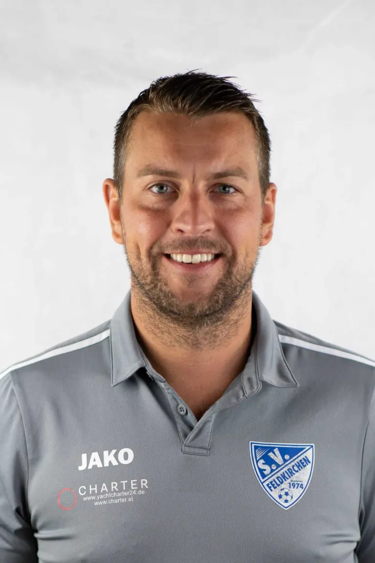 Ein Mann trägt ein graues Shirt mit weißen Streifen, dem Wort Jako in weißen Buchstaben und einem blauen Logo auf der linken Seite.