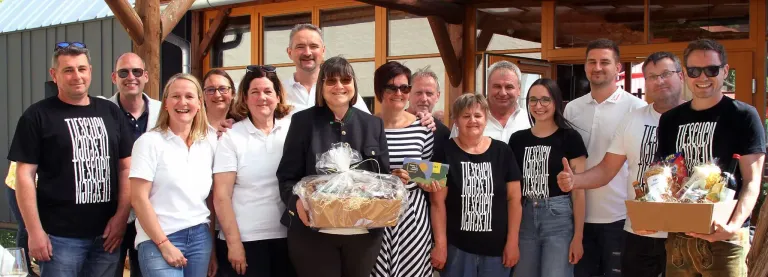 Eine Gruppe von Menschen steht draußen in der Nähe eines Gebäudes. Eine Frau hält einen Korb mit einem Geschenk darin.