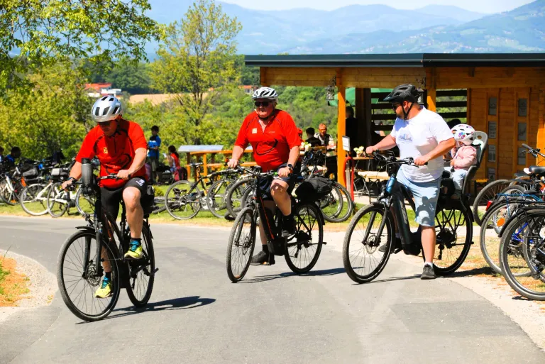 Three men ride bicycles on a road with a building and greenery behind them. They wear helmets and cycling attire. Mountains and more trees are in the distance.