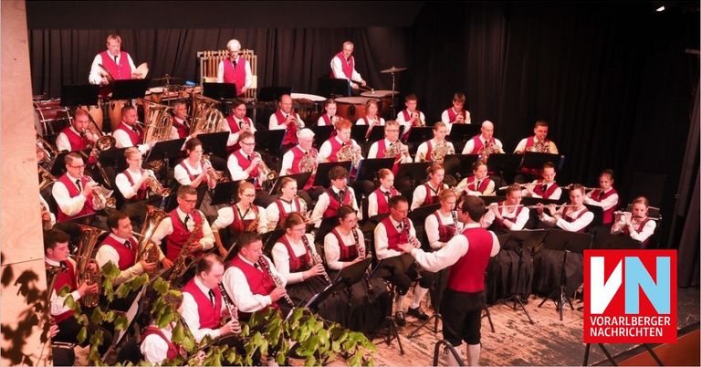 Eine Gruppe von Musikern in roten Westen und schwarzen Krawatten spielt auf der Bühne. Sie spielen verschiedene Instrumente wie Trompeten, Posaunen und Tuben. Die Bühne ist mit schwarzen Vorhängen im Hintergrund dekoriert.