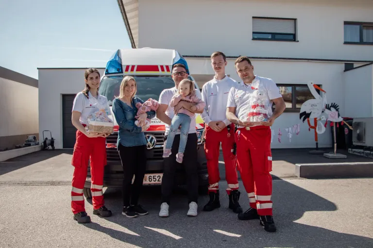 Fünf Personen stehen vor einem Krankenwagen, alle tragen rote Uniformen. Ein Mann hält ein Baby, die anderen haben Körbe. Sie lächeln für ein Foto.