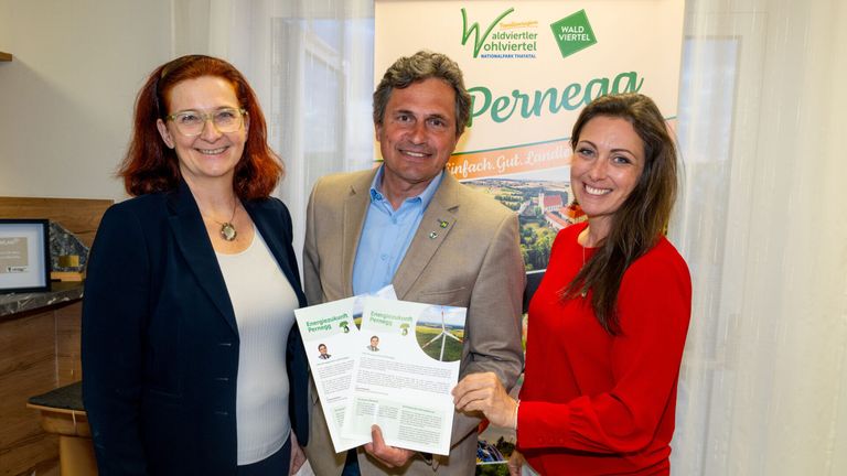 Drei Menschen posieren für ein Foto. Der Mann hält Papiere mit Bildern von Windrädern. Die Frauen tragen Brillen und eine Halskette. Hinter ihnen ist ein Poster mit grünem Text und einem Bild einer Landschaft.