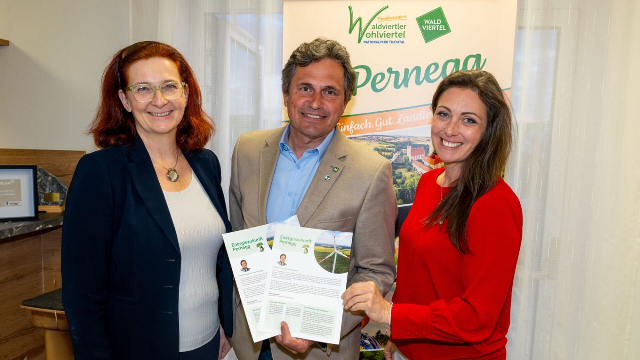 Drei Menschen posieren für ein Foto. Der Mann hält Papiere mit Bildern von Windrädern. Die Frauen tragen Brillen und eine Halskette. Hinter ihnen ist ein Poster mit grünem Text und einem Bild einer Landschaft.