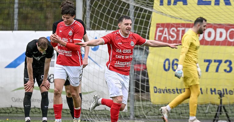 Fußballspieler feiern ein Tor bei einem Spiel. Ein Spieler in Rot trägt die Nummer 5, ein anderer in Weiß die Nummer 15. Ein gelbes Werbebanner ist im Hintergrund sichtbar.