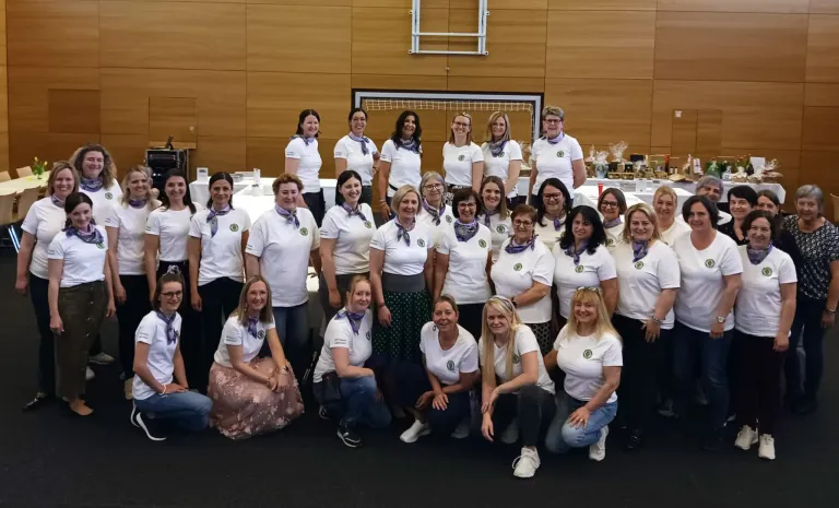 Eine Gruppe von Frauen in weißen Shirts und Schals posiert für ein Foto, einige sitzend und einige stehend in einem Raum mit einer Holzwand.